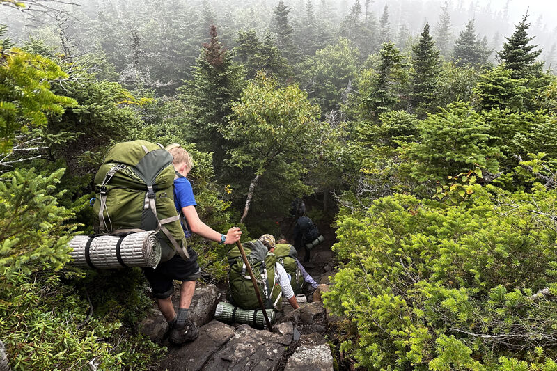 The image shows a group of hikers ascending a rocky, wooded trail. The foreground features a hiker with a large green backpack and a sleeping bag attached. They are using a hiking stick for support. The path winds upwards through dense green vegetation and trees, with mist or fog visible in the background, suggesting a high-altitude or mountainous environment. Other hikers are visible further up the trail.